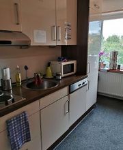 Eine kleine Küchenzeile mit beigen Schränken und dunkler Arbeitsplatte. Zu sehen sind ein Waschbecken, Herd, Mikrowelle und verschiedene Küchenutensilien. Rechts steht ein Wasserspender vor einem Fenster, durch das Bäume sichtbar sind. 