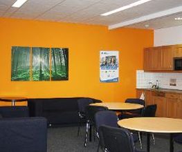Pausenbereich mit brauner Küchenzeile, runden Tischen und Stühlen sowie einem dunkelblauen Sofa und Sessel. Die Wand ist orangefarben und an der Wand hängt ein Leinwandbild von einem Wald.