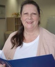 Eine Frau mit weißem Shirt und rosa Strickjacke hält lächelnd einen blauen Ordner in der Hand. Im Hintergrund sind unscharf ein Regal, ein Kühlschrank und ein Tisch mit Stühlen zu sehen. Der Raum wirkt hell und freundlich.