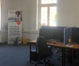 Schulungsraum mit mehreren Arbeitsplätzen jeweils mit 2 Bildschirmen, einem Bürostuhl, Tastatur, Maus und Headset. Im Hintergrund ein WBS TRAINING Roll Up Banner und eine Pflanze.