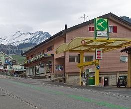 AGROLA Tankstelle in Amden. Gelbe Glasüberdachung mit einer Zanksäule und Zahlterminal. Digitaler Preisanzeige. Dahinter rotes Gebäude mit Volg und Bergkette im Hintergrund der Aufnahme.