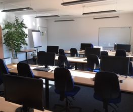 Schulungsraum mit mehreren Tischreihen an den blaue Bürostühle stehen. Auf den Tischen befinden sich Computerbildschirme sowie Tastatur und Maus.