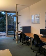 Schulungsraum mit mehreren Arbeitsbereichen, die durch blaue Trennwände voneinander abgegrenzt sind und mit 2 PC-Bildschirmen, Tastatur, Maus und Headset ausgestattet sind. Zugang zu einem Balkon.