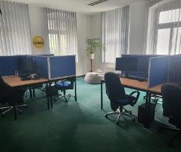 Schulungsraum mit mehreren Arbeitsplätzen. Jeweils mit 2 Bildschirmen, einem Sessel, Tastatur und Maus. In der Ecke steht eine Pflanze hinter einem Sitzsack.