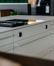 Die Detailaufnahme zeigt eine hochwertige Kücheninsel mit eleganten weißen Auszügen, schlanken Designgriffen und einer modernen Arbeitsplatte in Betonoptik. Im Mittelpunkt steht ein flächenbündig integriertes Induktionskochfeld mit Muldenlüfter, ergänzt durch eine Grillplatte für vielseitige Kochmöglichkeiten. Die Kombination aus klaren Linien, hochwertigen Materialien und durchdachter Funktionalität macht diese LEICHT Designküche zu einem Highlight moderner Küchenplanung im Küchentreff Liebold.