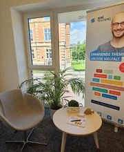 Sitzbereich mit einem Sessel und einem Beistelltisch auf dem Deko steht. Im Hintergrund ein Roll Up Banner und eine Zimmerpflanze vor dem Bodentiefen Fenster