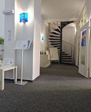 Empfangsbereich mit links einem Beistelltisch und Plakaten. Im Hintergrund ein Regal neben einer Wendeltreppe.