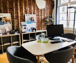 Modern und gemütlich gestalteter Beratungsraum. Weißer Tisch, graue Lounge-Sessel. Vor der Backsteinwand flache Regale mit Infomaterial. Großes Sprossenfenster. Parkettfußboden.