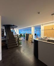 mediaService Bremen | Full-Stack Werbeagentur - Blick in das großzügige, offene Foyer einer Agentur mit dem Empfangstresen, einer dunklen Treppe in das Obergeschoss und angrenzenden Büroarbeitsplätzen.