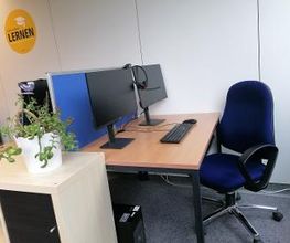 Arbeitsplatz bestehend aus 2 PC-Bildschirmen, Tastatur, Maus und Headset sowie einem blauen Bürostuhl und einer blauen Trennwand.