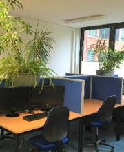 Mehrere Arbeitsplätze neben einem Fenster. Auf dem Tisch sind zwei Monitore und eine Tastatur zu sehen.