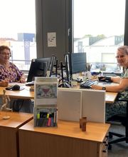2 Mitarbeiterinnen sitzen in ihrem Büro an ihren Schreibtischen vor ihren Computerbildschirmen. Im Hintergrund ein geöffnetes Fenster mit WBS TRAINING Logo Spiegelung.