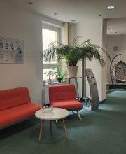 Eingangsbereich mit grünem Teppichboden. Links ein rotes Sofa mit Beistelltisch, dahinter eine Pflanze und Flyerständer. Im Hintergrund ein Hängesessel und ein TV.