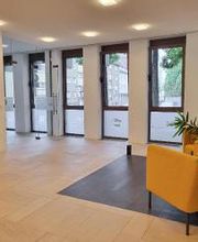 Heller Wartebereich mit Fensterfront und 2 orangenen Sessel. Im Vordergrund steht eine große Zimmerpflanze.