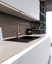 Diese elegante LEICHT Designküche überzeugt mit einer puristischen Gestaltung, hochwertigen Materialien und einer durchdachten Lichtinszenierung. Die weiße, grifflose Arbeitszeile wird durch eine schwarze Einbauspüle und eine edle Edelstahlarmatur stilvoll ergänzt. Indirekte LED-Beleuchtung unter den Oberschränken sorgt für eine angenehme Arbeitsatmosphäre und setzt die klaren Linien der Küche gekonnt in Szene. Offene Designregale mit schwarzen Metallrahmen verleihen der modernen Wohnküche zusätzliche Leichtigkeit und bieten Platz für stilvolle Dekoration.