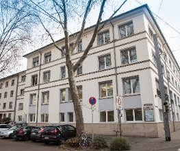 Seitliche Außenansicht auf das Gebäude von der gegenüberliegenden Straßenseite. Hier wird die Ansicht von der Hauptstraße auf das seitliche Gebäude präsentiert. Bäume und Parkmöglichkeiten.