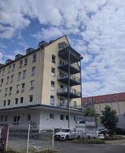 gelbes Gebäude mit vielen Fenstern und Balkonen an der Seite. Unten steht eine Tafel mit Beschilderungen und ein paar Autos.