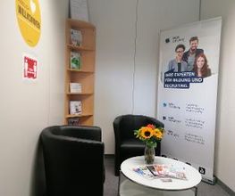 Gemütliche Sitzecke mit 2 schwarzen Ledersesseln, einem weißen Beistelltisch auf dem ein Blumenstrauß steht sowie ein Werbeaufsteller und Holzregal im Hintergrund.