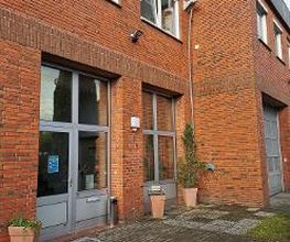 Ein Backsteingebäude mit großen Fenstern und einer Glastür. Vor dem Eingang stehen Blumenkübel mit Pflanzen. Rechts ist ein großes Garagentor zu sehen. Der Boden vor dem Gebäude besteht aus Pflastersteinen und einem kleinen Bereich mit Gras.