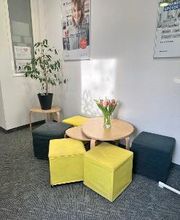 Sitzbereich mit 5 Sitzhockern und einem Beistelltisch auf dem Tulpen stehen. Dahinter eine Topfpflanze und Plakate an der Wand.