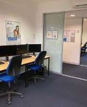 Offener Lernbereich mit vier PC-Arbeitsplätzen mit jeweils 2 Bildschirmen, einem Bürostuhl, Tastatur, Maus und Headset. An der Wand eine Uhr und ein Plakat.