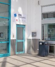 Außenansicht vom Eingangsbereich. Türkise Tür- und Fensterumrahmung. Über der Eingangstüre hängt ein WBS Training Schild mit der Aufschrift „Unser Herz schlägt für Bildung“.