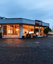 Die Außenansicht zeigt das Küchenstudio Küchentreff Liebold in den Abendstunden. Große Glasfronten geben den Blick auf die stilvoll beleuchtete Küchenausstellung frei und präsentieren moderne Designküchen in hochwertigem Ambiente. Die einladende Architektur, das markante Küchentreff-Logo und die helle Innenbeleuchtung vermitteln Kompetenz, Qualität und Inspiration für individuelle Küchenplanung.