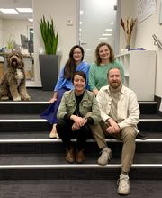 Grruppenbild vom Standortteam. Alle sitzen auf einer Treppe und lachen freundlich in die Kamera. Daneben steht ein Hund.