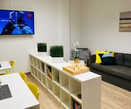 Modern und hell gestalteter Aufenthaltsbereich mit zwei weißen Tischen und gelben Stühlen. Rechts ein dunkelgraues Sofa mit Kissen. Zwischen den Tischen und dem Sofa Ikea-Kallax Raumteiler. Blumendeko, Gesellschaftsspiele, Bücher im Regal. Wandbild.