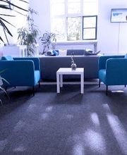 Heller Aufenthaltsraum mit 2 blauen Sesseln und weißen Beistelltisch, 2 graue Sofas und einem Fernseher an der Wand. Im Raum stehen einige, grüne Zimmerpflanzen.