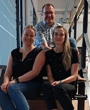Ein Teil des Standortteams sitzt im Flur auf einer Treppe und lächelt fröhlich in die Kamera. 2 Frauen und ein Mann.
