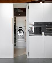 Moderne, grifflose Hochschrankküche mit integrierter Geheimtür zu einem praktischen Hauswirtschaftsraum. Hinter der flächenbündigen Schrankfront verbergen sich Waschmaschine, Stauraum und Arbeitsbereich – eine elegante Lösung für ein aufgeräumtes, funktionales Wohnkonzept.