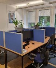 Arbeitsplätze mit blauen Trennwänden, 2 Bildschirmen, Tastatur, Maus und Headset. Der Raum hat 3 Fenster mit Blick ins Grüne. An der Wand hängen Bilder von Personen.
