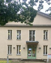 Außenansicht vom Gebäude. Helle Fassade mit weißen Fensterrahmen und einer grünen Eingangstür.