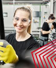 Eine Mitarbeiterin von  actuell Persona arbeitet konzentriert an einer Maschine in einer modernen Produktionshalle. Mit Schutzbrille und Arbeitshandschuhen steht sie für Sicherheit, Präzision und professionelles Arbeiten in der Industrie. Im Hintergrund sind weitere Mitarbeitende bei technischen Tätigkeiten zu sehen, was Teamarbeit und den praxisnahen Arbeitsalltag unterstreicht.