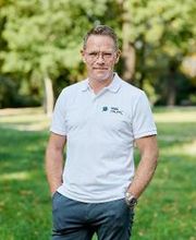 Stefan Schrader steht im weißen T-Shirt mit WBS GRUPPE Logo in der grünen Natur und lächelt in die Kamera.