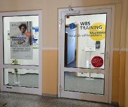 Eingangsbereich mit zwei Glastüren. Auf der Linken ist ein WBS-Poster aufgeklebt, auf der rechten steht "WBS TRAINING. Herzlich willkommen"