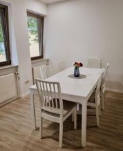 Weißer Tisch mit 6 Stühlen im Pausenraum. Auf dem Tisch steht eine Vase mit Blumen.