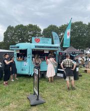 FirstLoveCoffee GmbH - Kaffee Catering Berlin - Mobiler Kaffeeanhänger auf grünem Festivalgelände mit Bio-Kaffee, Iced Latte, Iced Chai, Fahnen und Barista-Service für Sommer-Events.