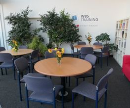 Gemütlicher Aufenthaltsraum mit runden Tischen, auf denen Blumen stehen, grünen Zimmerpflanzen, Bücherregal und rotem Sofa. Auf der Wand steht WBS TRAINING – unser Herz schlägt für Bildung geschrieben.
