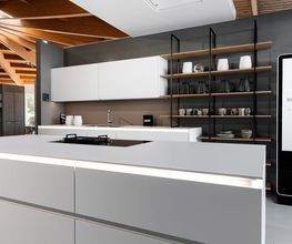 Moderne, grifflose Designküche mit großzügiger weißer Kücheninsel, integriertem Kochfeld und indirekter LED-Beleuchtung. Offene Holzregale mit schwarzem Metallrahmen, eine dunkle Rückwand und die sichtbare Holzbalkendecke schaffen einen eleganten Kontrast zwischen minimalistischem Küchendesign und natürlicher Architektur.