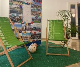 Foyer mit Fußballdekoration. 2 Liegestühle auf einem grünen Teppich und 2 Fußbälle. Dahinter ein Aufsteller mit verschiedenen Fotos und eine Zimmerpflanze.