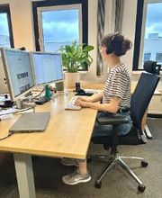 Arbeitsplatz mit 2 Bildschirmen, Tastatur und Maus. Eine Frau sitzt auf einem Bürostuhl am Tisch und arbeitet. Im Hintergrund steht eine Zimmerpflanze.