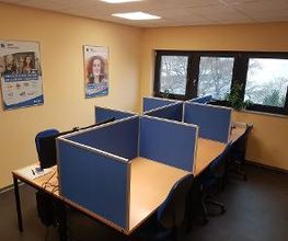 Schulungsraum mit mehreren Arbeitsbereichen, die durch blaue Trennwände voneinander abgegrenzt sind und mit PC-Bildschirmen, Tastatur, Maus und Headset ausgestattet sind. Fensterreihe und Bilder an der Wand.