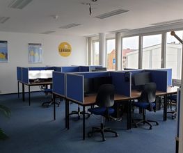 Schulungsraum mit mehreren Arbeitsbereichen, die durch blaue Trennwände voneinander abgegrenzt sind und mit 2 PC-Bildschirmen, Tastatur, Maus und Headset ausgestattet sind sowie blauen Bürostühlen.