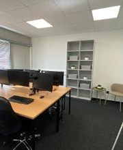 Schreibtische im Trainingsraum mit jeweils zwei PC-Bildschirmen, Tastatur, Maus und Headset. Im Hintergrund steht ein Bücherregal. Daneben ist eine Sitzgelegenheit mit einem Beistelltisch