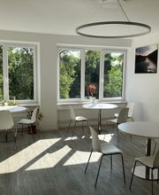 Ein heller Aufenthaltsraum mit großen Fenstern mit Blick ins Grüne. In dem Raum befinden sich runde, weiße Tische und passende Stühle. An der Wand hängen Leinwandbilder von Hamburg.
