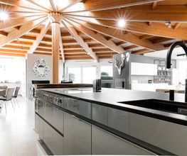 Diese exklusive Designküche verbindet moderne Eleganz mit natürlicher Architektur. Im Vordergrund stehen eine großzügige Kochinsel mit integriertem Bora Kochfeld, eine mattschwarze Quooker Design-Armatur und ein flächenbündiges Spülbecken. Die offene Raumgestaltung mit beeindruckender Holzbalkendecke, hochwertigen Materialien und klaren Linien schafft ein stilvolles Wohnambiente. Die hochwertige LEICHT Küche fügt sich harmonisch in den lichtdurchfluteten Wohnraum ein und vereint Funktionalität mit zeitlosem Küchendesign.