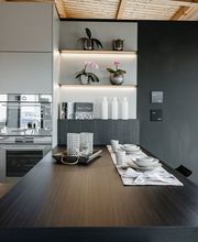 Moderne Designküche mit großzügiger Kochinsel und integrierter Sitzgelegenheit. Die Kombination aus matten Fronten, dunkler Holzoptik, offenen Regalen mit indirekter LED-Beleuchtung und stilvoll gedecktem Esstisch schafft eine elegante und wohnliche Atmosphäre. Hochwertige Einbaugeräte sowie durchdachte Stauraumlösungen machen diese LEICHT-Küche zu einem perfekten Beispiel für modernes Küchendesign.