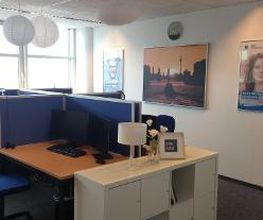 Raum mit mehreren Arbeitsplätzen mit jeweils 2 Bildschirmen, einem Bürostuhl, Tastatur und Maus. Ein weißes Regal mit Lampe steht am Kopf der Arbeitsplätze. An der Wand hängt ein großes Bild und 2 Plakate.